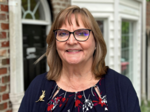 Leslee Sharp, a white woman with light brown hair, wears a navy shirt with red and white flowers, a navy sweater, and a gold kangaroo pin on her sweater.