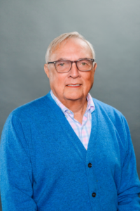 Leo, a white man with white hair and brown glasses, wears a pink and pale blue plaid shirt and blue button-down sweater.