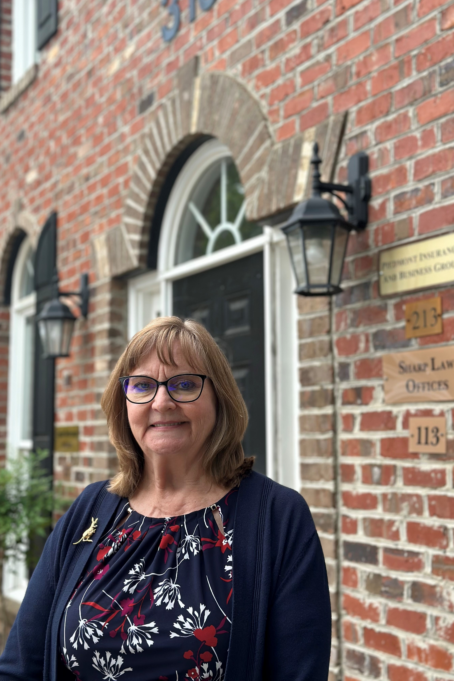 light brown hair, wears a navy shirt with red and white flowers, a navy sweater, and a gold kangaroo pin on her sweater. She stands outside her office, which is in a red brick building and has a gold sign with "Sharp Law Offices" outside the black door.