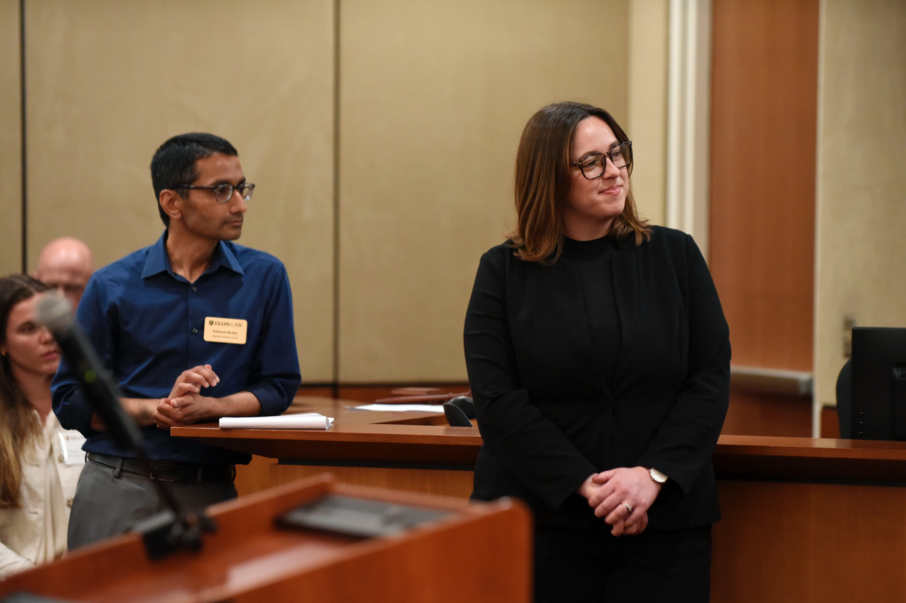 Megan, a white woman with shoulder-length brown hair and brown glasses, wears a black blouse and suit. She stands with a man, a professor named Srikanth Reddy, who has black hair and black glasses and is wearing a blue button-down shirt and grey pants.