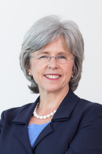 Rosemary, a white woman with grey hair and clear glasses, wears a blue blouse, black jacket and white necklace. 