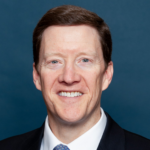 Jim, a white man with brown hair, wears a white shirt, blue tie and black jacket.