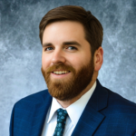 Kris, a white man with brown hair and a beard, wears a white shirt, blue tie and dark blue jacket. 