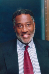 Charles, a Black man with black hair and a beard, wears a blue shirt, red tie and navy suit.