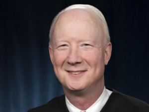 Judge Bledsoe, a white man with white hair, wears a white shirt and black judge's robe.