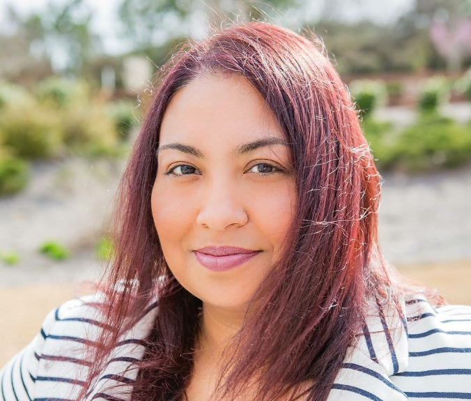 Elysia, a woman with auburn hair, wears a white blouse and white and navy blue striped blazer.