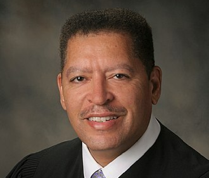 Judge Wynn is a Black man with black hair. He wears a white shirt, purple tie and black judge's robe.