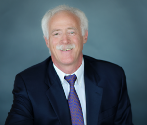 Kerry, a white man with white hair and a mustache, wears a white shirt, purple tie, navy jacket and khaki pants.