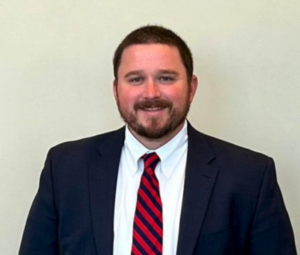 Tripp, a white man with brown hair, wears a white shirt, red and navy striped tie, and navy suit.