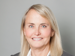 Paula, a white woman with blond hair, wears a white blouse and grey blazer.