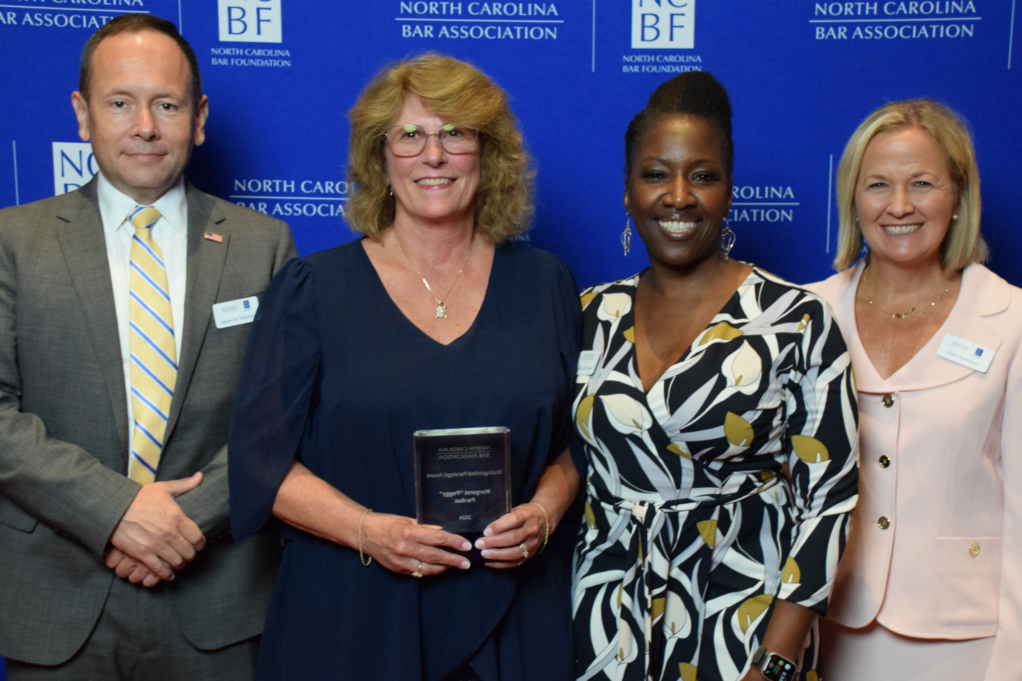 Peggy, a white woman with light brown hair and glasses, wears a navy blue dress and a silver necklace. She is pictured with Jason Hensley, Lakisha Chichester, and Patti Ramseur.