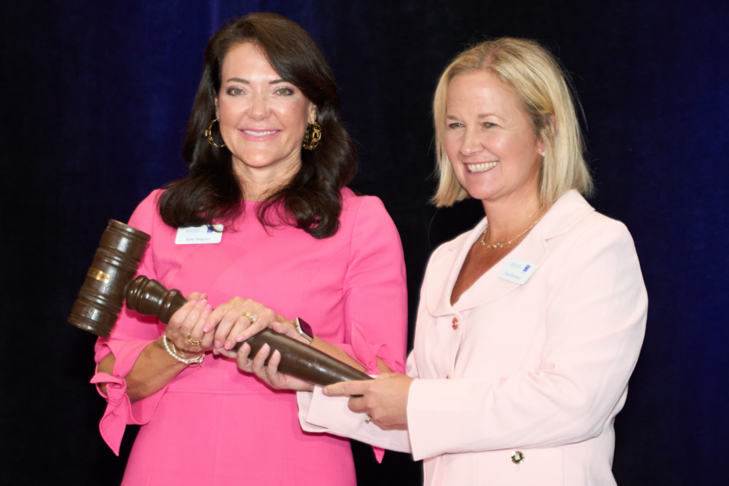 Kim Stogner, a white woman with dark brown hair, wears gold earrings and a bright pink dress. She and past president Patti Ramseur, a white woman with blond hair who wears a pale pink suit, hold the brown gavel and are pictured smiling.