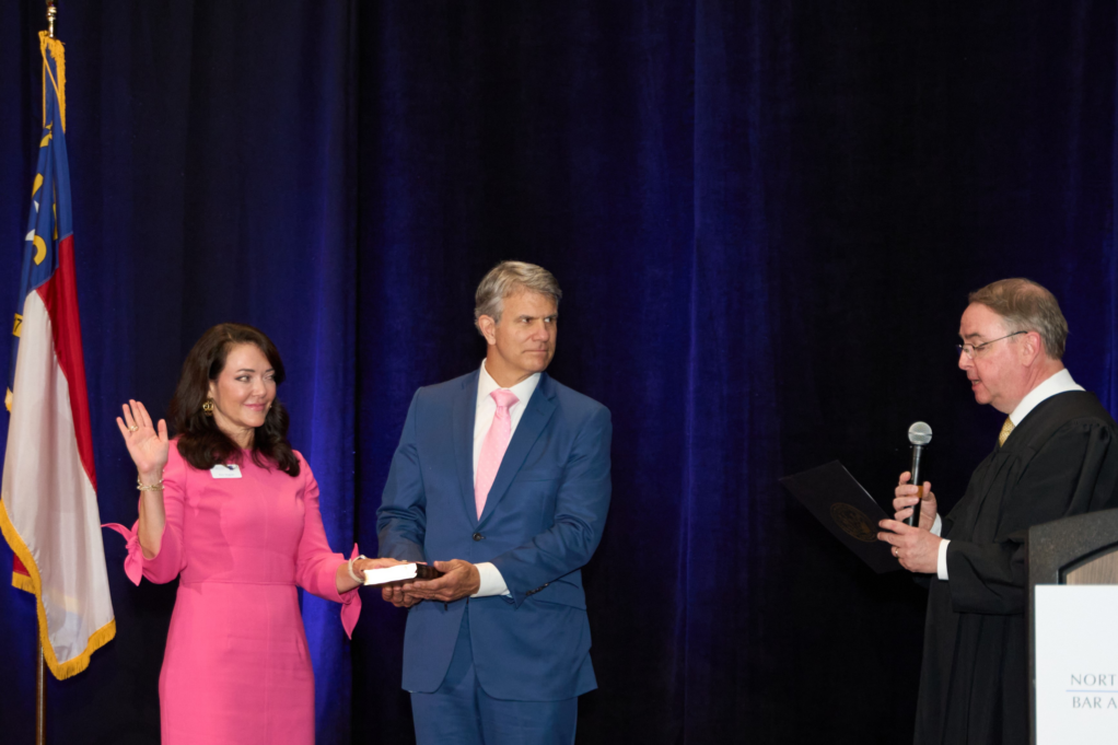 Kim Stogner, a white woman with dark brown hair, wears a bright pink dress. She has her hand on a Bible, and she is standing next to her husband, Stuart, who wears a white shirt, pink tie, and blue suit. Stuart holds the Bible as Superior Court Judge Eric C. Morgan administered the oath of office.