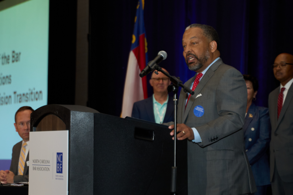 Rob Harrington is pictured speaking at a podium. Rob is a Black man with black hair and a beard and is wearing a blue shirt, red tie and grey suit.