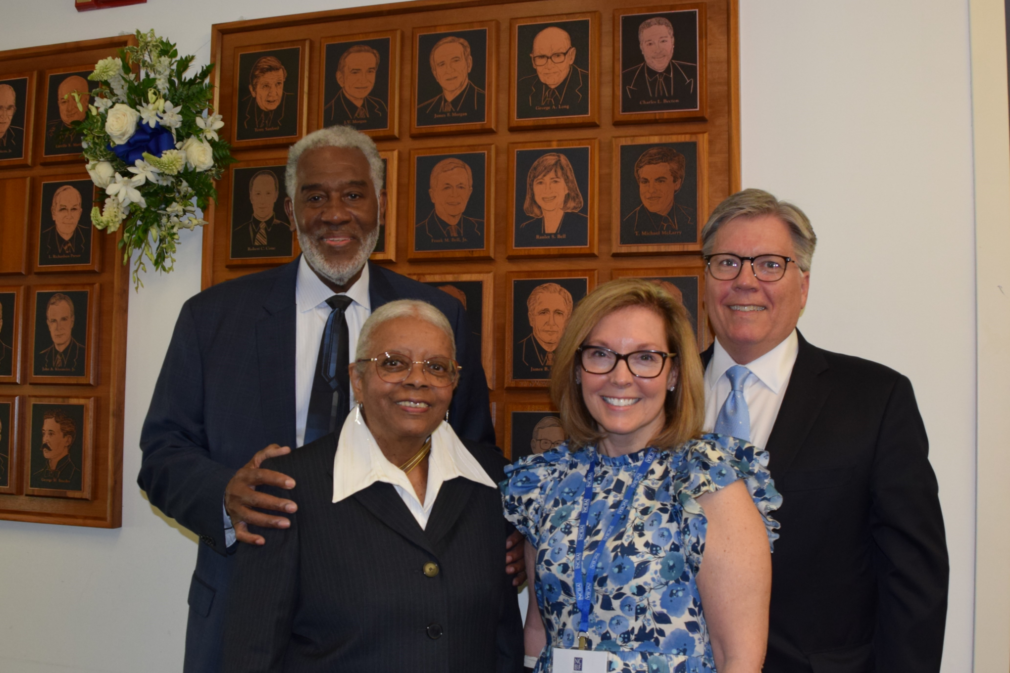 Charles Becton is a Black man with white hair wearing a white shirt, black tie, and black suit, and he stands with his hand on his wife's shoulder. Brenda Becton is a Black woman with white hair wearing a white shirt and black suit. Mark Holt stands near his wife, Joanna. Mark is a white man with brown hair wearing a white shirt, blue tie and black suit, and Joanna is a white woman with brown glasses wearing a white dress with blue flowers.