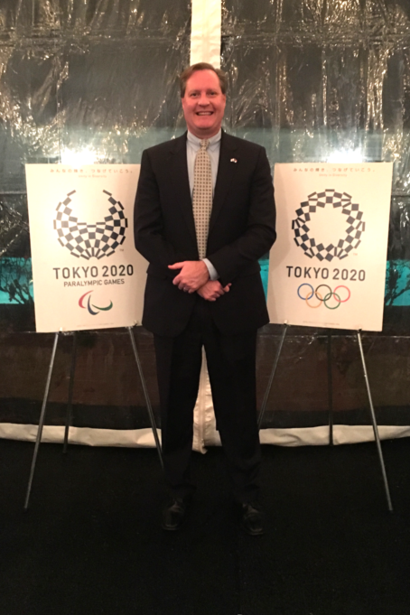 Robinson, a white man with brown hair, wears a grey shirt, beige tie and black suit, and he is photographed between two signs. One reads "Tokyo 2020 Paralympic Games" and "Tokyo 2020" with the Olympic logo.