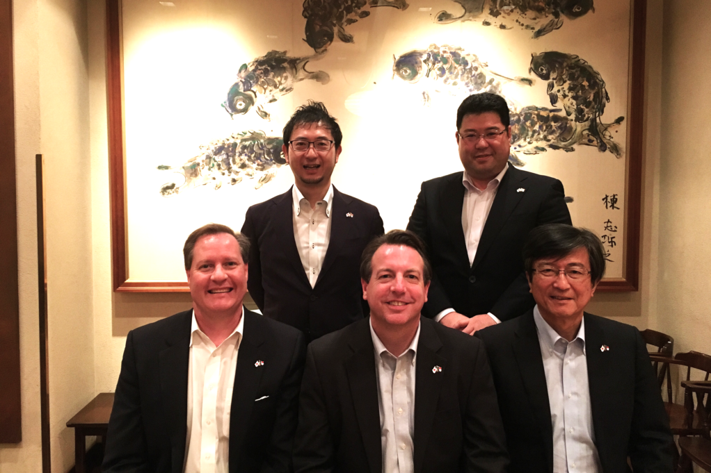 Robinson, a white man with brown hair who is wearing a white button-down shirt and suit, is seated with two other individuals. Three members of the Daini Bar Association are pictured in a dimly lit restaurant.