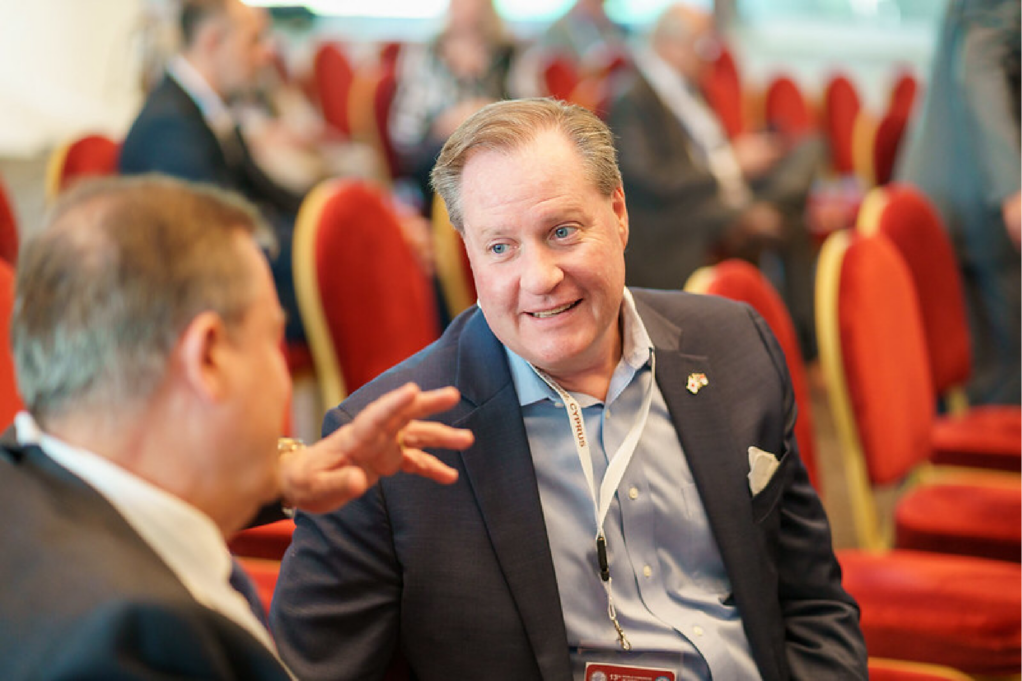 Robinson, a white man with brown hair, wears a grey button-down shirt and black suit. He is seated smiling and talking with another man and there are red chairs with people in the background.