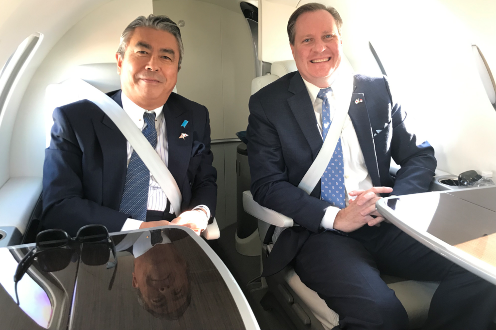 Ambassador Sugiyama and Robinson are pictured flying in a HondaJet. The ambassador wears a white shirt, blue tie and navy blue suit, and Robinson wears a white shirt, blue tie with white print and navy suit. They are pictured smiling.