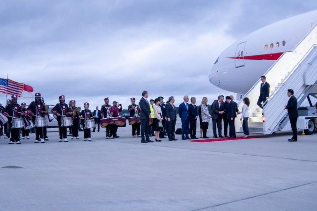 The photo shows the Prime Minister's plane on the tarmac at RDU airport. The Prime Minister is standing on a red carpet and Governor Cooper, a white man with grey hair, and other people are greeting him.