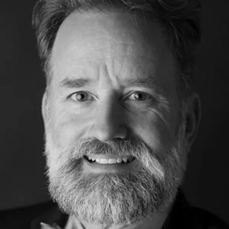 John, a white man with brown hair and a beard, is pictured in black and white and is wearing a light shirt with a dark blazer.