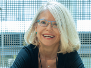 Mary Craven Adams, a white woman with blond hair and teal glasses, wears a black blouse, teal earrings and a necklace with a white gem. She is pictured smiling.