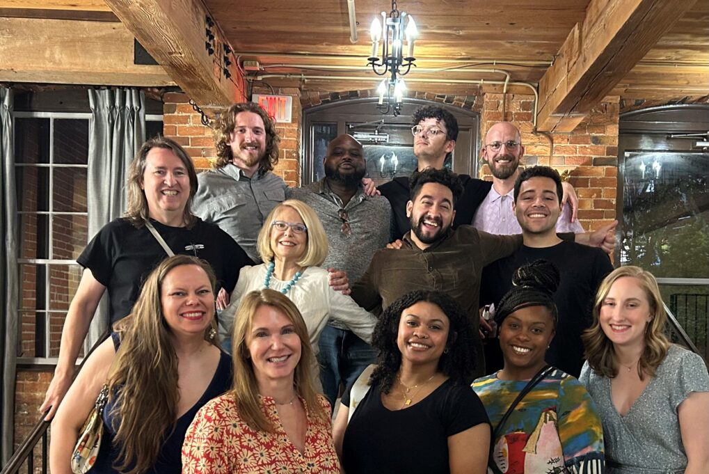 Mary is pictured with the 11 members of her graduating cohort. Mary wears a white blouse and teal necklace. Everyone is smiling. They are pictured with a brick wall and windows behind them.