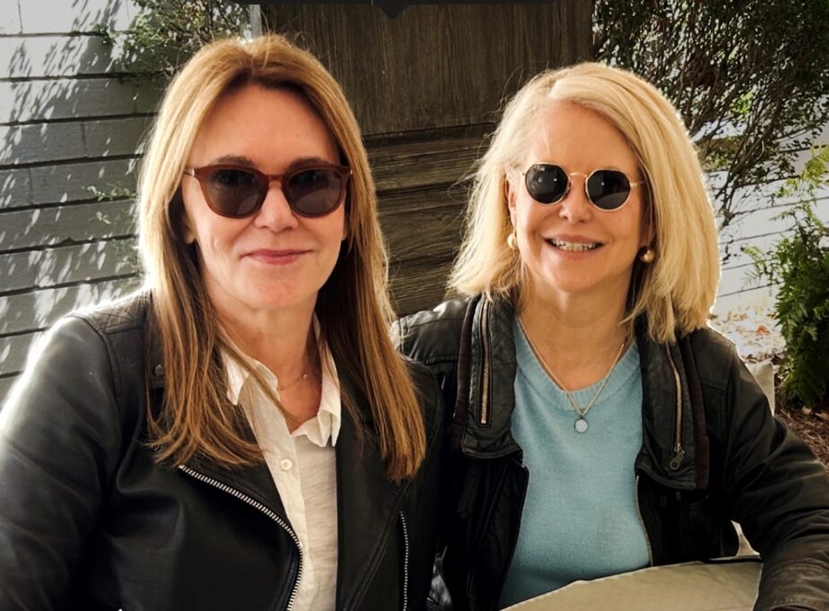 Mary, a white woman with blond hair, wears sunglasses, a teal t-shirt and black jacket. Agatha is a white woman with brown hair who wears a white t-shirt and a black jacket.