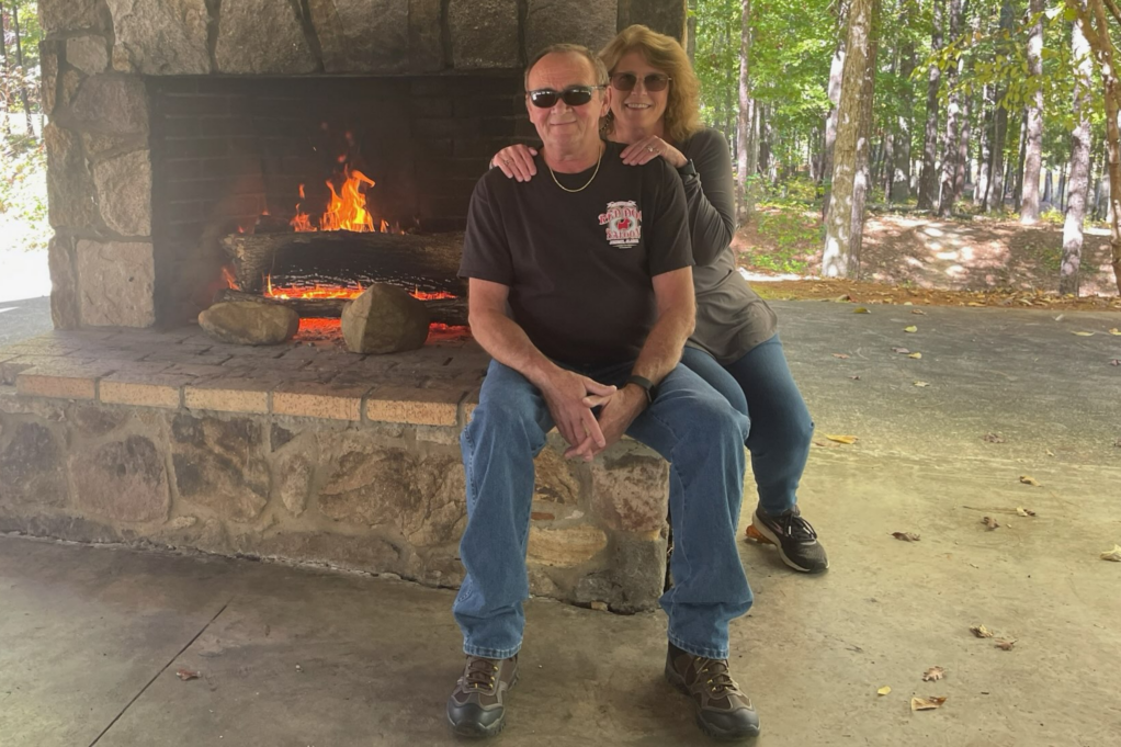 Peggy Pardue, a woman with light brown hair, wears jeans and a green sweater. She and her husband, a man with brown hair wearing a black shirt and jeans, are pictured sitting on an outdoor fireplace with trees in the background.