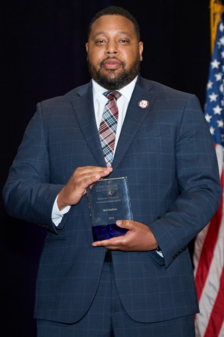 William Walton is a Black man with black hair and a beard. He is wearing a white shirt, a red and burgundy plaid tie, and a blue suit with light blue stripes. He is pictured holding his award.