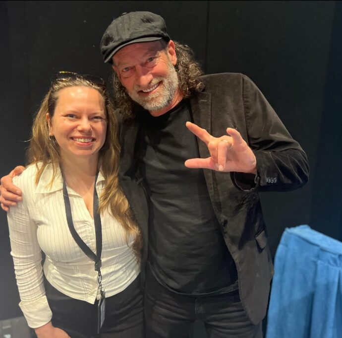 Autumn, a white woman with long brown hair, wears a white blouse. Troy, a white man with brown hair and a beard, wears a black t-shirt, pants, jacket and black hat. 