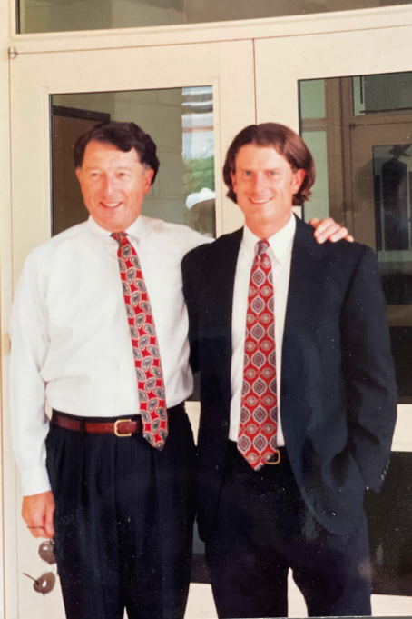 Coleman, a white man with brown hair, wears a white shirt, black suit and red tie. His father, Don, stands beside him with his arm on his shoulder. Don is a white man with brown hair and is wearing a white shirt, red tie, and black pants.