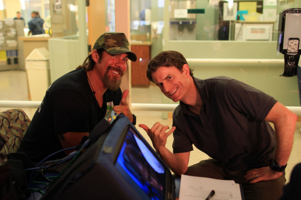 Cowan, a white man with brown hair, wears a black polo, and is seated next to Tim Medvetz, a white man with brown hair and a beard who is wearing a camo colored hat and black t-shirt.