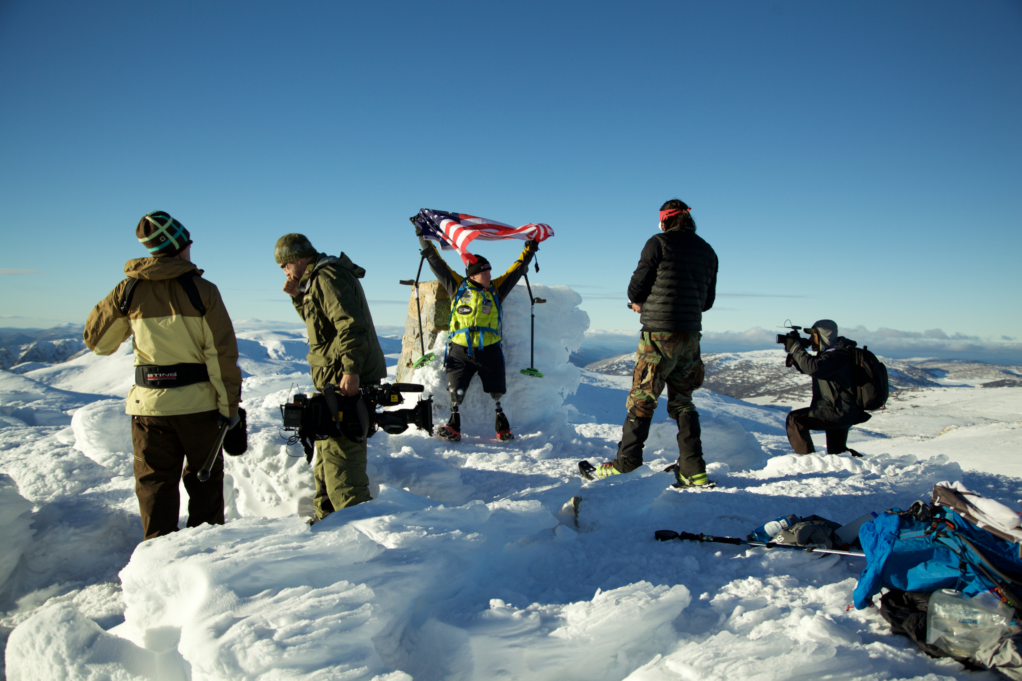 Sergeant Blunt is pictured at the peak of the moutain. He wears a bright green vest, black jacket, and black pants. He raises the American flag above his head and four members of the 60 Minutes crew are pictured around him.