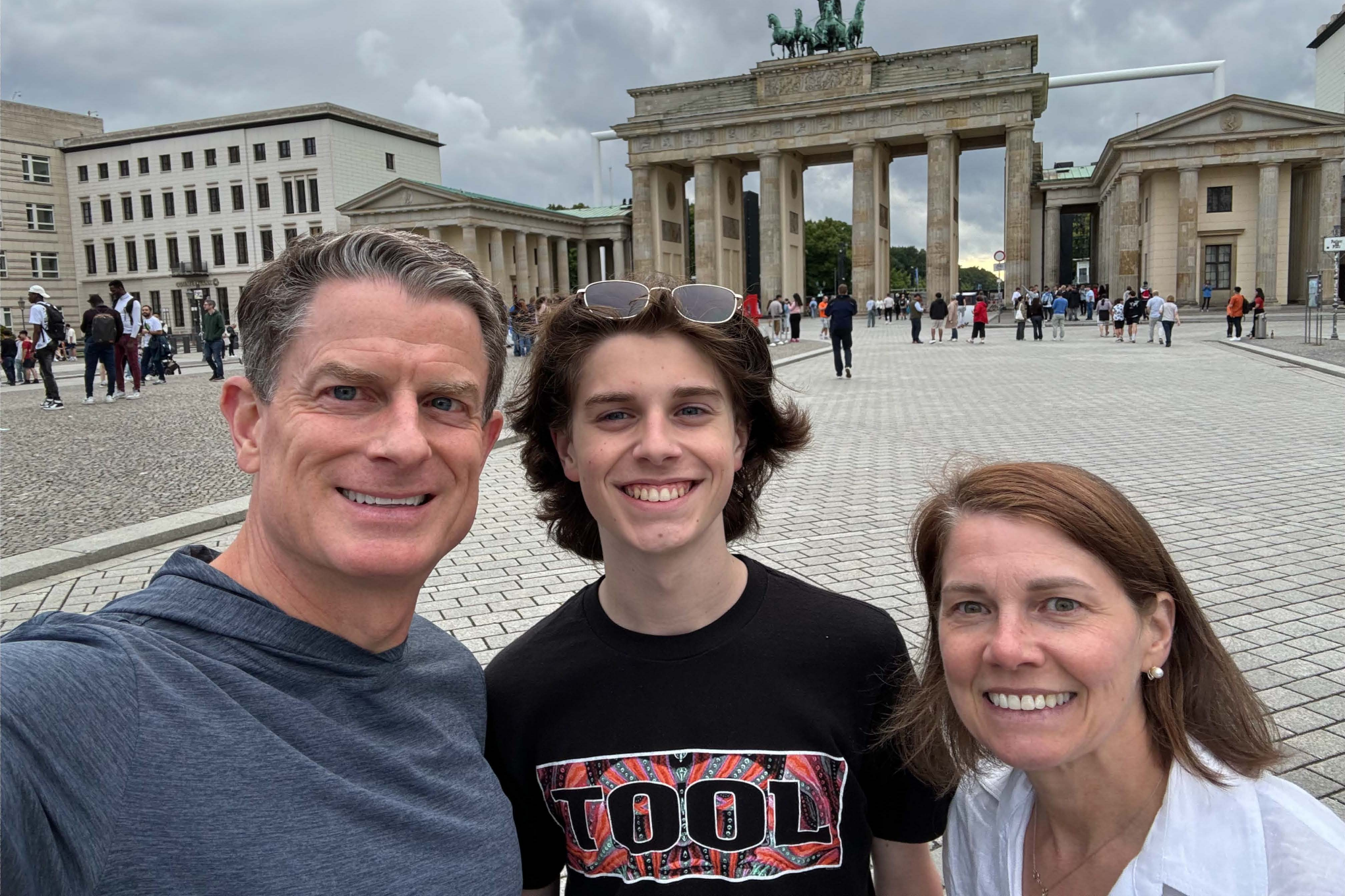 Coleman, a white man with brown hair, wears a grey hoodie. His son Julian, a white young man with brown hair and wearing a black shirt stands in the middle. Angie, a white woman with brown hair, wears a white blouse and pearl earrings. They are in an open area with historical buildings behind them.