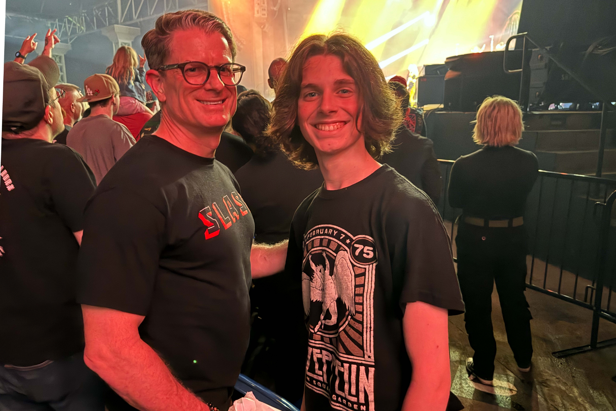 Cowan, a white man with brown hair and brown glasses, wears a black shirt, and Julian, a white young man with brown hair, wears a black Led Zeppelin t-shirt. You can see a crowd and stage behind them.