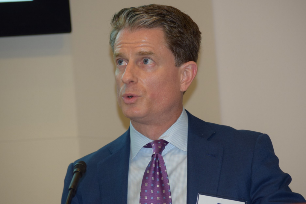 Cowan, a white man brown hair and blue eyes, is pictured speaking at a CLE. He wears a pale blue shirt, purple tie and navy suit.