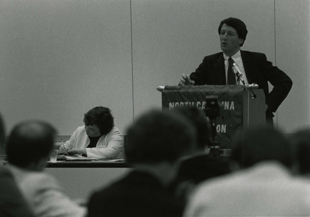 Don, a white man with dark hair, wears a suit and tie and stands at a podium speaking. The photo is in black and white.