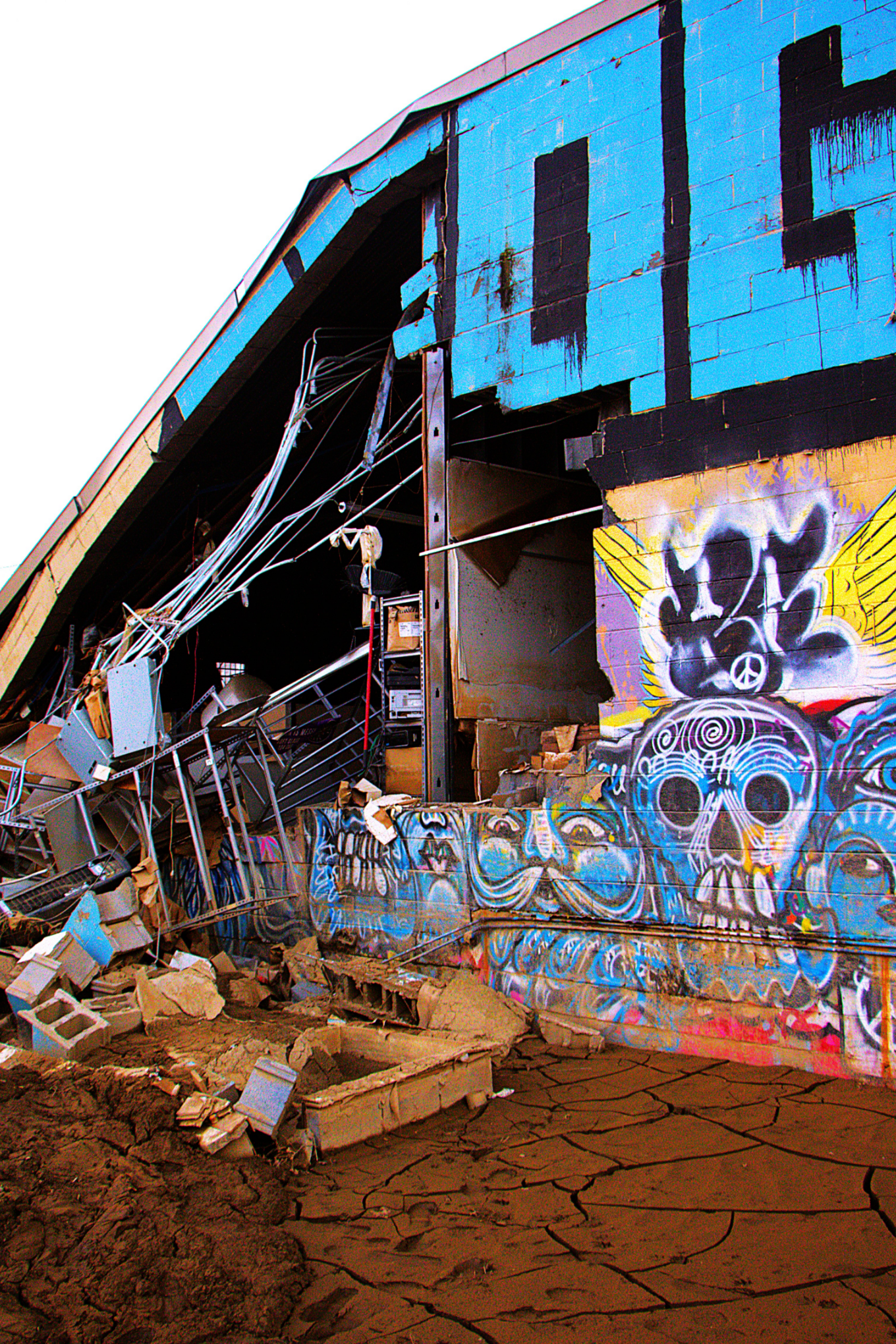 A bright blue building with graffiti on it has collapsed on one side, with wood and other debris below it. 