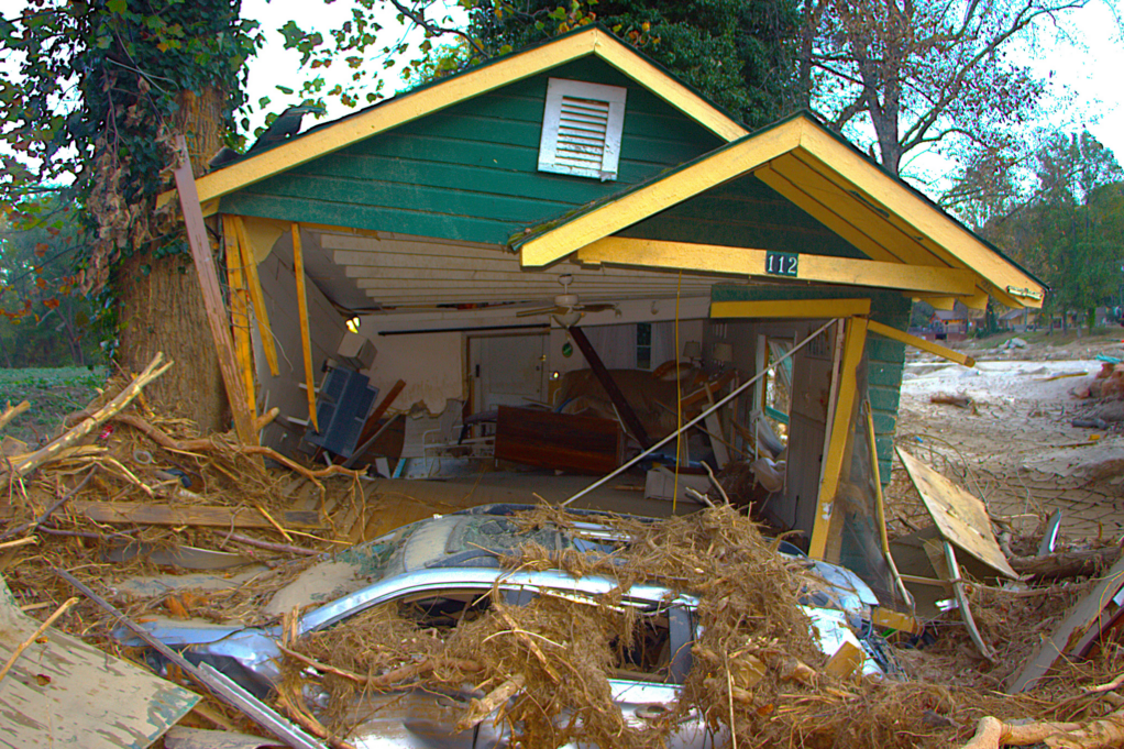 Storm damage from Helene is shown in this photo. A small green house has been destroyed, and a car with debris and trees on it is shown in front of the house.